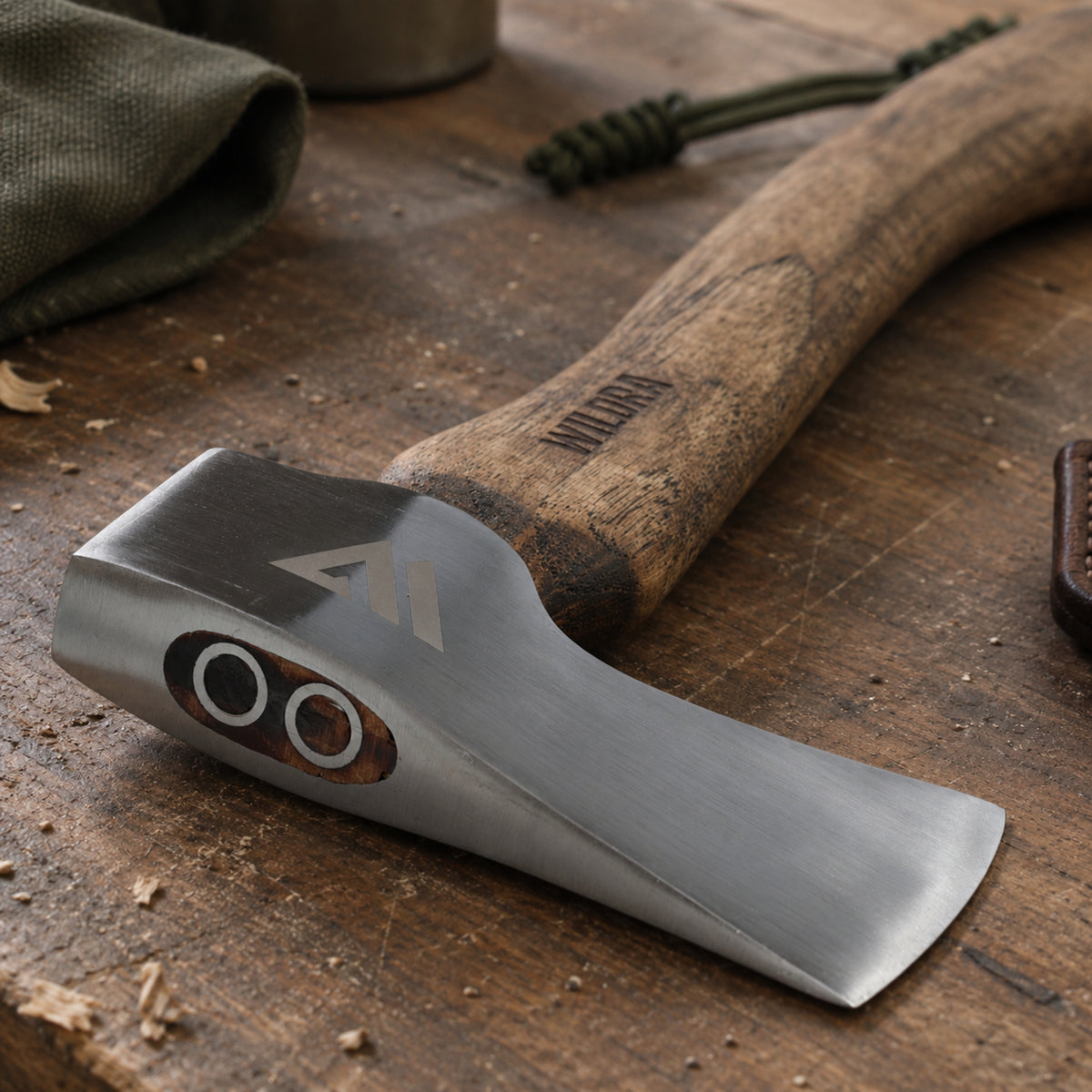 A close-up of a hand axe or hatchet with a brushed silver steel head and a brown wooden handle, resting on a rustic wooden surface. The head has a stamped geometric logo, and the handle is engraved with the brand name 'MikroLondo'.