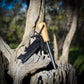 Multitool with wooden handle and black strap attached to a tree branch in a natural setting