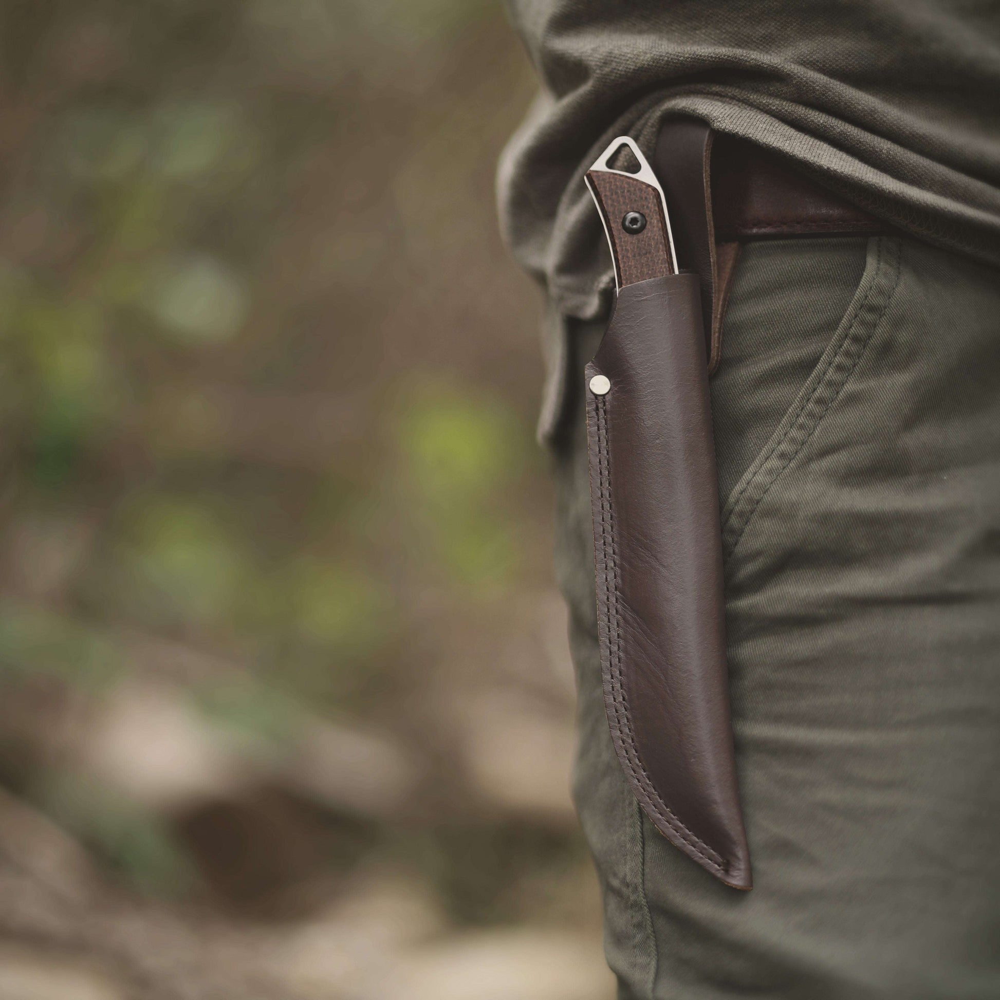 Knife with leather sheath attached to a person's pocket against a blurred natural background